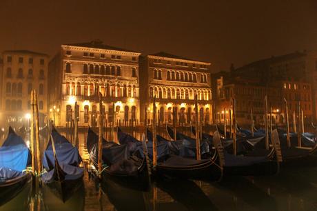 A foggy evening scene with a row of gondolas docked in front of beautifully lit historic buildings, their warm glow reflecting off the water in Venice.