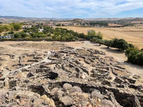 The ancient ruins of a Nuragic village in Sardinia, with circular stone structures scattered across the dry landscape, surrounded by distant fields and olive trees.