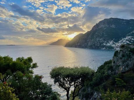 A stunning coastal view on the Amalfi Coast at sunset, with the sun's golden rays illuminating the sea and the rugged cliffs, with lush green trees framing the scene and a coastal town visible in the distance.