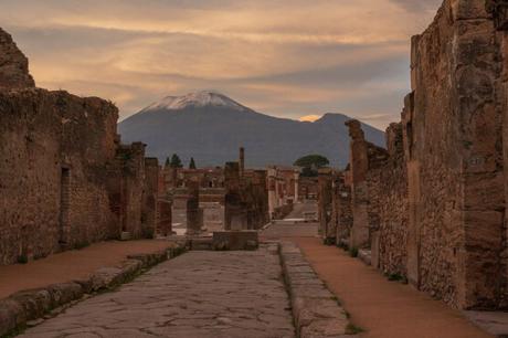 A cobblestone street lined with ancient ruins in Pompeii, with a distant view of a snow-covered Mount Vesuvius under a golden sky.