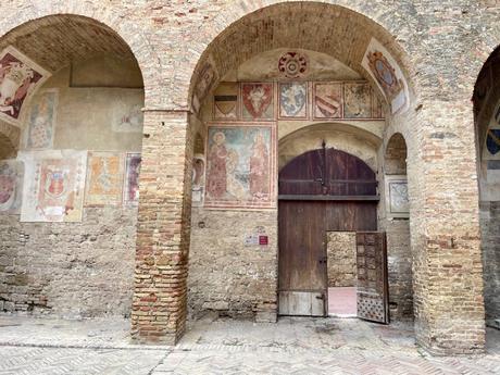 A detailed view of a medieval-style brick archway and walls decorated with faded frescoes, depicting various coats of arms and religious figures, inside a historic building.