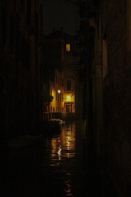 A dark, narrow canal in Venice illuminated by a single lit window and glowing street lights, with boats floating quietly in the water.