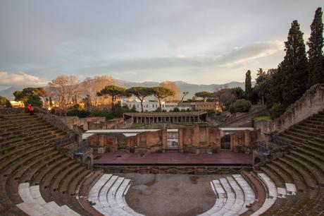 The grand amphitheater in Pompeii, with rows of stone seating and brick ruins, surrounded by trees and mountains in the distance.