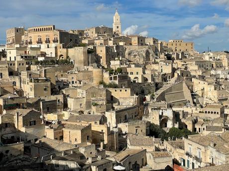 A view of the center of the ancient hilltop city of Matera, Italy with tightly packed stone buildings with beige facades, stacked on different levels, creating a picturesque and historic urban landscape under a clear sky.