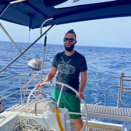 Alex Tiffany smiling and steering a sailboat on a sunny day, with a backdrop of open blue ocean and a clear sky.