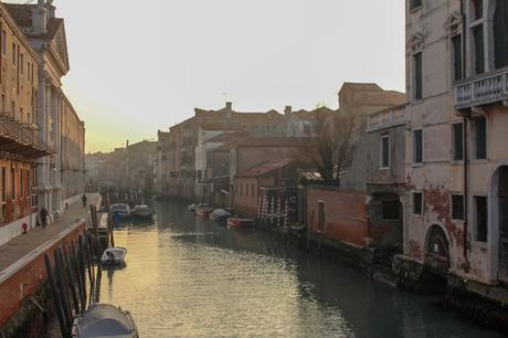 A wide Venetian canal at sunrise with historic buildings lining the water, bathed in soft, golden light.