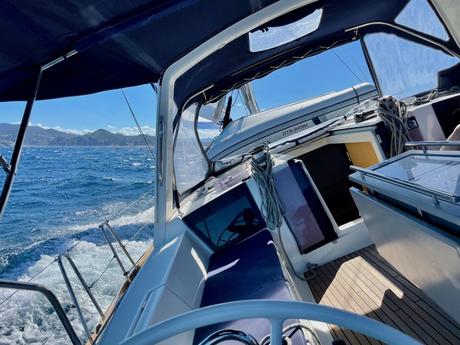 A view from a boat’s cockpit as it sails through open water, with rugged mountains visible on the horizon, under a clear blue sky.