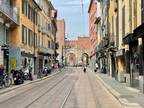 A street scene in Milan, Italy, with tram tracks running through a narrow road lined with colorful buildings and scooters parked along the sidewalks. In the distance, an archway is visible.