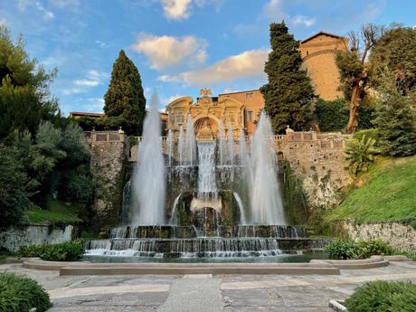 A majestic fountain in Tivoli's Villa d'Este, with large water jets shooting up from multiple levels surrounded by greenery and historical architecture, under a soft blue sky with clouds.