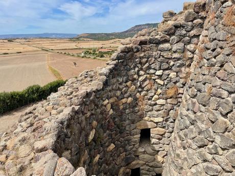 A close-up view of stone walls forming the remains of a Nuragic tower, with views of rolling farmland and hills stretching into the distance.