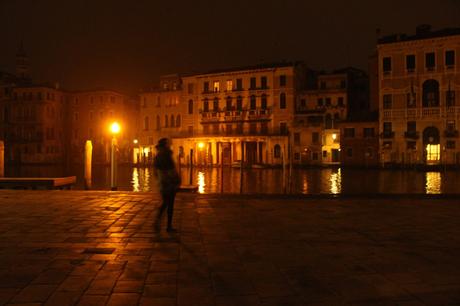 A blurry figure walking on a Venetian square at night, with dimly lit buildings along the canal reflecting light on the water.