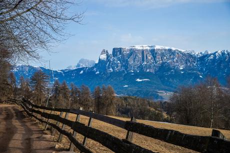 A scenic view of snow-capped mountains in the Dolomites, framed by a rustic wooden fence and a bare tree, with patches of snow visible in the valley below.