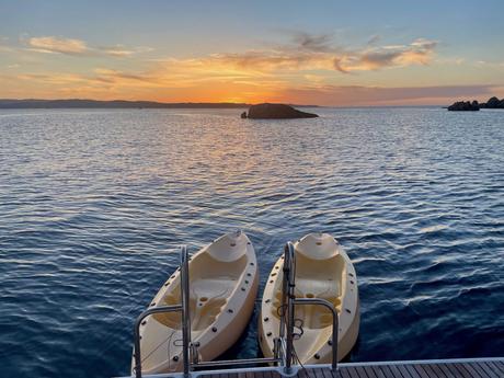 A tranquil sunset over the sea with two yellow kayaks tied to the back of a larger boat, the water gently reflecting the orange hues of the setting sun.