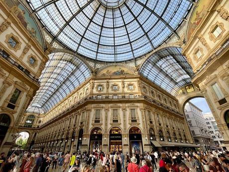 A grand indoor shopping gallery in Milan with an ornate glass dome ceiling, intricate architecture, and a crowd of people walking and browsing stores, including luxury brands like Louis Vuitton.