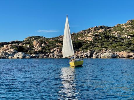 A bright yellow sailboat on calm waters, surrounded by rocky hills and lush greenery under a clear sky.