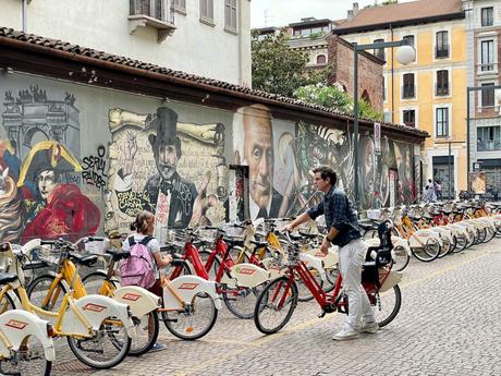 A colorful mural on a wall in Milan, featuring historic figures like Napoleon, with rows of parked bicycles and a man helping a child with a bike.