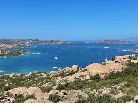 A panoramic view of a coastal area on the Costa Smeralda in northern Sardinia with deep blue water and scattered sailboats. The shoreline is dotted with rocky formations and greenery.