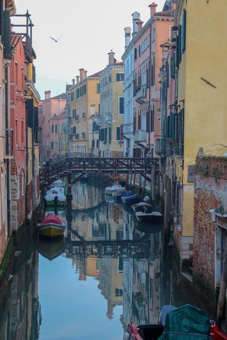 A serene Venetian canal with brightly colored buildings reflected on the water, and small boats moored alongside a wooden bridge.