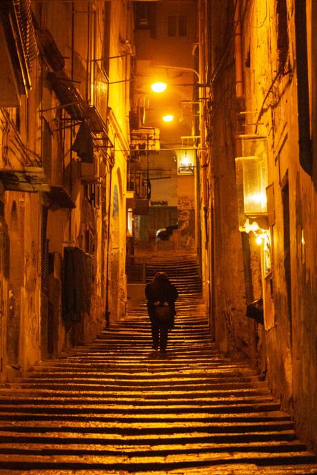 A dimly lit, narrow street in Italy at night, with orange streetlights illuminating the steps and a lone person walking up the stairs.