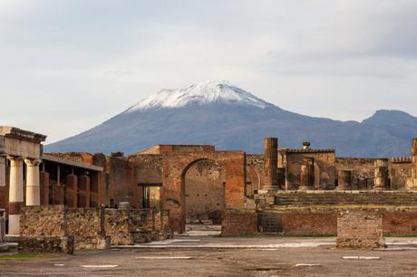 The ancient ruins of Pompeii, with a snow-capped Mount Vesuvius towering in the background under a cloudy sky.