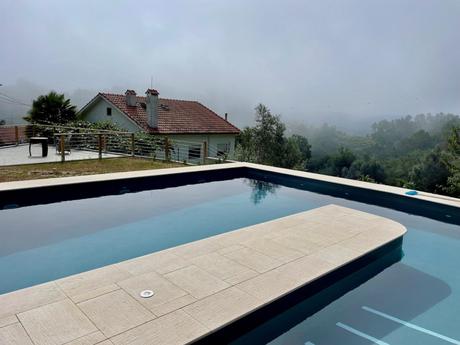 A tranquil backyard pool in northern Portugal surrounded by greenery, with a house in the background and misty hills visible in the distance. The calm water of the pool reflects the foggy sky, creating a serene atmosphere.