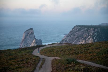 A scenic view of a rocky coastline with a towering rock formation rising out of the water. A single figure walks down a dirt path through the grassy cliffs, with the expansive ocean stretching out in the distance.