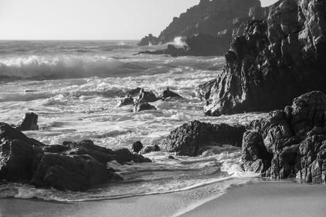 A dramatic black-and-white image of a rocky shoreline with waves crashing over large boulders. The texture of the rocks and the swirling ocean create a striking contrast, highlighting the natural beauty of the rugged coastline.