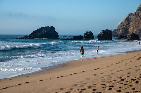 A serene beach scene with a woman walking along the shoreline as gentle waves roll in. Large, jagged rocks rise from the ocean in the distance, while a few other beachgoers enjoy the water near the rocky shore.