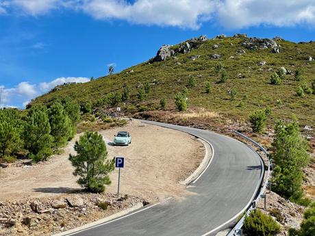 A small green Fiat 500 car parked on a dirt pullout next to a winding road that curves around a green hillside. Sparse trees dot the rugged landscape, and the sky above is bright blue with a few clouds.