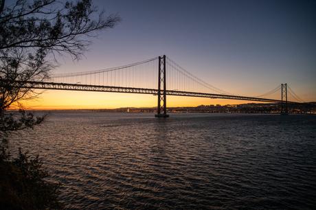 A large suspension bridge stretches across the calm Tagus River during sunset, with the sky transitioning from a warm orange near the horizon to a deep blue above. Tree branches frame the scene on the left, and city lights can be seen glowing faintly in the background.