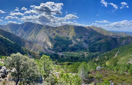 A panoramic view of lush green mountains, with valleys dotted with small villages and farmland. The mountains rise steeply against a clear blue sky filled with puffy white clouds.