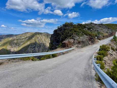 A scenic mountain road cuts through rocky terrain, with a metal guardrail along the side. The surrounding hills are covered in green vegetation, and a few fluffy clouds dot the bright blue sky.