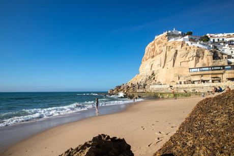 A peaceful beach scene at Azenhas do Mar, Portugal with soft waves crashing against the golden sand. In the background, a towering cliff is topped with white buildings, and a few people walk along the shore, enjoying the sunny day under a clear blue sky.