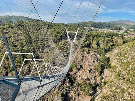 The metal 516 Arouca Bridge stretches across a deep rocky gorge, with lush green forests covering the surrounding cliffs. The intricate cables of the suspension bridge create a dramatic pattern against the clear sky.