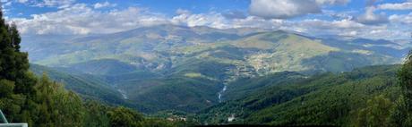 A panoramic view of rolling green mountains under a bright blue sky with scattered clouds. A river winds through the valley below, surrounded by dense forests and a few distant houses.