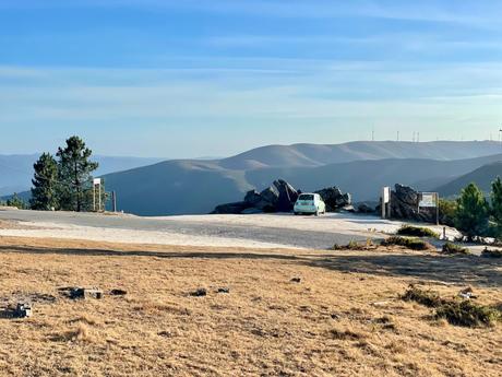 A small green car parked at a scenic overlook, with rolling hills and wind turbines visible in the distance. The barren, rocky terrain contrasts with the soft curves of the distant hills under a clear sky.