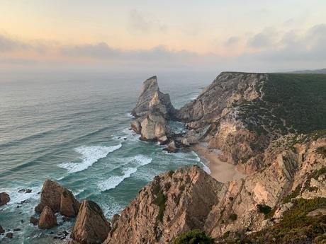 A dramatic cliffside landscape at sunset, with waves crashing against the rocks below. The ocean sparkles under the fading sunlight, and the rugged cliffs create a sense of vastness and isolation.