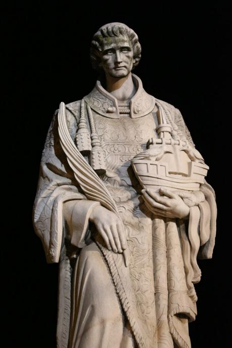 A marble statue of a solemn-looking figure dressed in ornate robes, holding a palm branch in one hand and a detailed model of a ship in the other. The statue is set against a dark background, emphasizing its intricate design and craftsmanship.