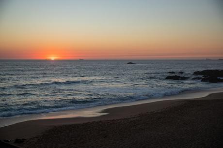 A beautiful sunset over the ocean, with the sun just dipping below the horizon near Sintra in Portugal. Soft waves lap against the sandy beach, and a few distant ships are visible on the horizon as the sky glows in shades of orange and pink.