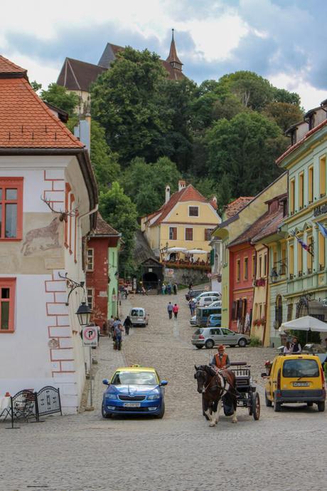 A lively street scene in a Romanian town, with cobblestone streets, colorful buildings, and a mix of cars and a horse-drawn carriage. People are walking up the hill towards the church on the hilltop.