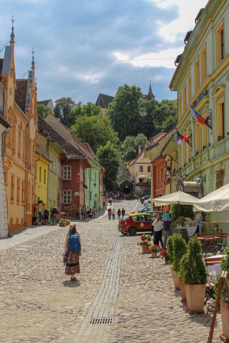 A cobblestone street in a Romanian town lined with colorful buildings on both sides. A person with a blue backpack is walking away from the camera, and the street leads uphill towards a church surrounded by greenery.