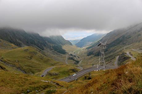 A wide view of a mountainous valley, partially covered in mist, with the winding Transfăgărășan highway snaking through the landscape. A tall electricity tower is visible on the right side of the image.