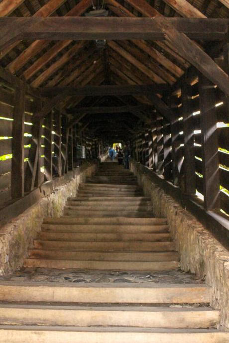 A covered wooden staircase with stone sidewalls, enclosed by dark wood beams and panels, leading upwards to a dimly lit destination, with a few people visible at the top.