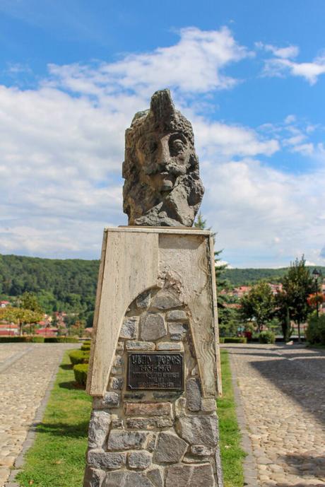 A stone bust of Vlad Țepeș, also known as Vlad the Impaler, set atop a monument with a plaque. The background shows green trees and a blue sky with some clouds.