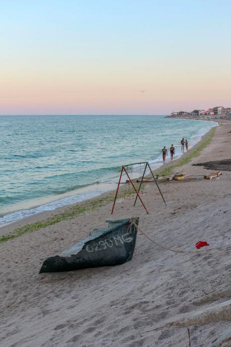 A beach scene at sunset in Vama Veche, with soft pastel hues in the sky. A broken swing set and a small boat are on the shore, while three people walk along the waterline in the distance.