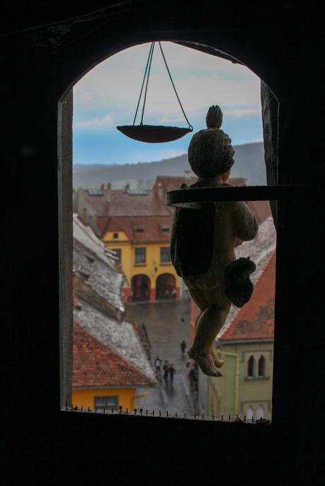 A view through a stone window of a small statue silhouetted against a medieval Romanian town. The streets and rooftops below are visible as people walk through the square.