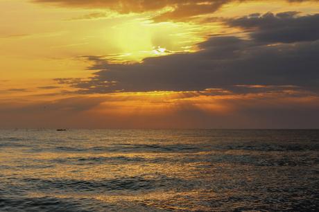 A serene view of the ocean at sunrise, with golden light streaming through the clouds. Seabirds fly over the water, and small fishing boats can be seen in the distance.