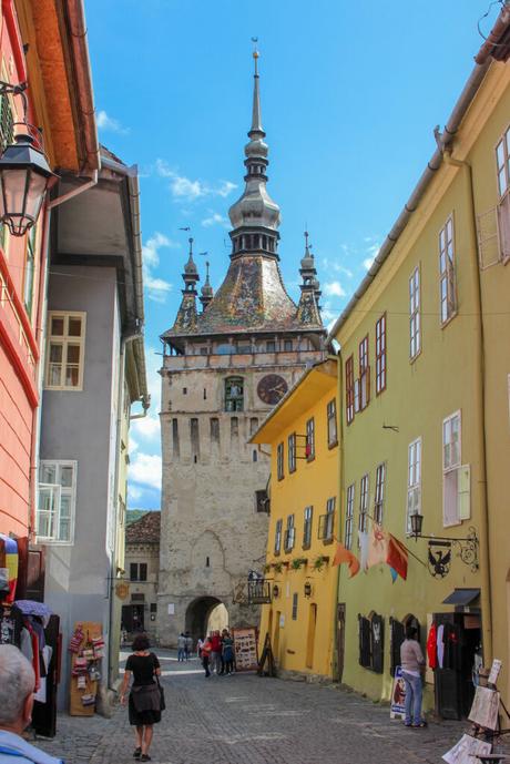 A picturesque street in Romania with a tall clock tower featuring spires in the distance. The buildings are painted in bright colors, with shops and pedestrians walking along the narrow cobblestone road.