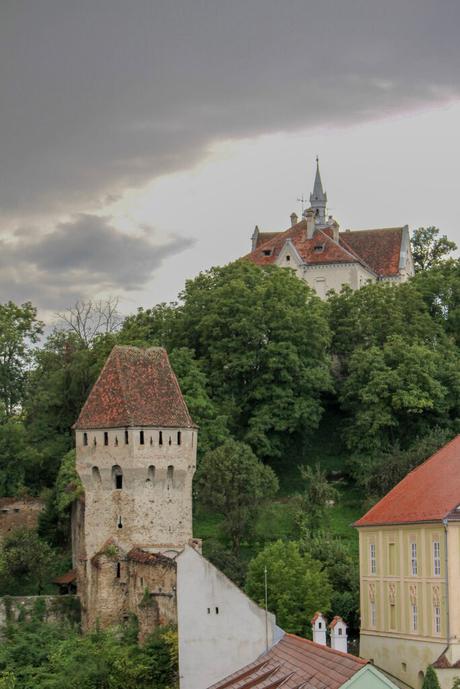 Portrait view from the Sighisoara Clock Tower, looking over the red-tiled rooftops and pastel-colored buildings of the medieval town. The lush green hills and distant landscape provide a scenic backdrop, encapsulating the charm of this Transylvanian village.