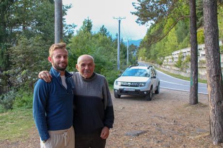 Alex Tiffany standing with an older Romanian man beside a road, with a white SUV parked in the background. The scene is surrounded by trees and mountains.